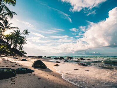 Plage tropicale avec des palmiers et des rochers, baignée par une lumière de soleil perçante, sous un ciel bleu partiellement nuageux