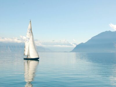 Voilier solitaire naviguant paisiblement sur des eaux calmes avec des montagnes enveloppées de nuages en arrière-plan.